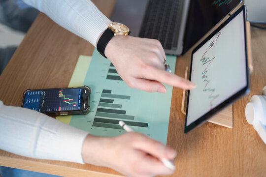 A person analyzes financial charts on a tablet and phone, with a laptop and bar graph on a wooden desk.