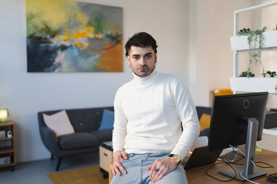A young man in a white turtleneck sweater and jeans sits on a desk in a modern office, looking at the camera.