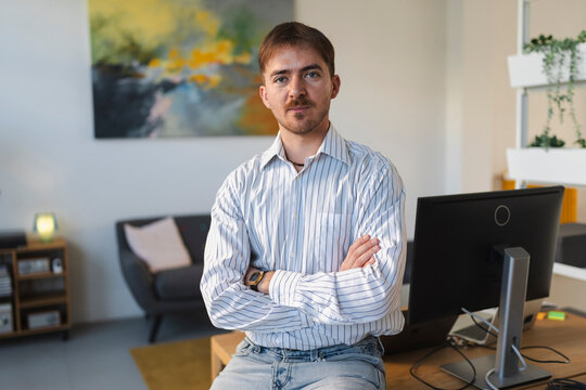 A man with crossed arms sits in an office, looking at the camera. He wears a striped shirt and jeans, with a computer and artwork in the background.