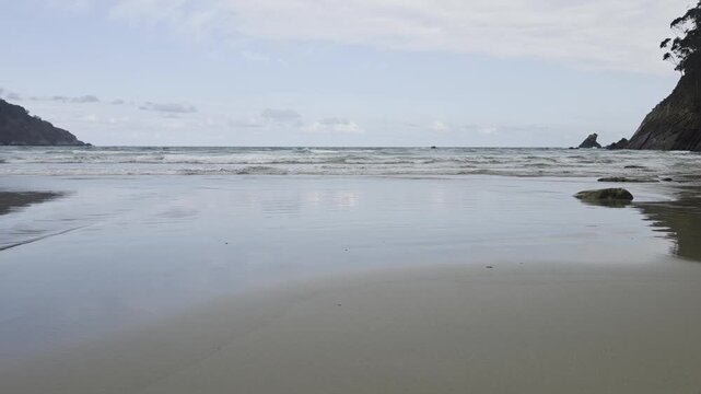 Static shot of empty sandy beach with gentle waves rolling onto rocky coastline under cloudy sky. Concha de Artedo, Spain