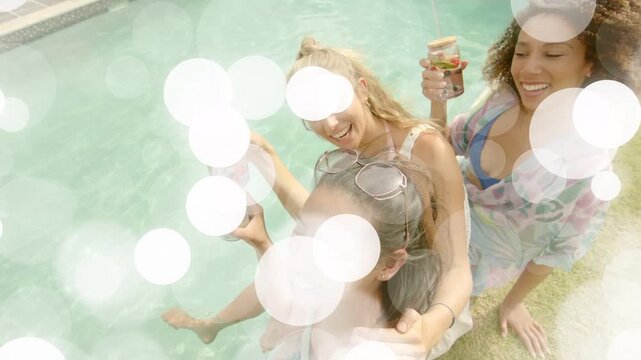 3 women toasting poolside as middle woman lifting drink while top-down camera adding bokeh for ads