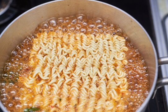 This dynamic close up captures traditional Korean spicy ramen boiling vigorously in a stainless steel pot. The bubbling red broth and curly wheat noodles perfectly highlight this comforting K-food.