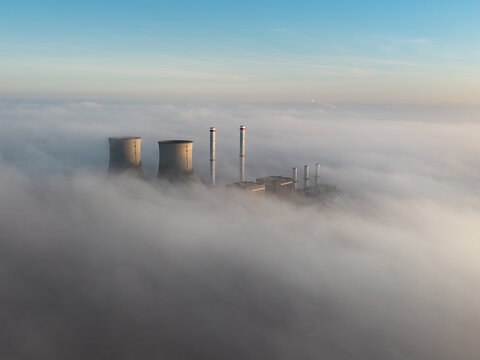 Aerial view of the RWE Clauscentrale power plant with cooling towers and chimneys emerging from a thick layer of morning fog under a clear blue sky in Maasbracht, Limburg, Netherlands.