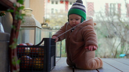 Baby kneeling in bear teddy fleece coat and striped beanie hat leaning on crate of apples, toddler exploring objects curiously during outdoor family day in cold season © Marco