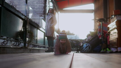 Baby crawling outdoors in bear teddy fleece coat and striped beanie hat with mother in beige coat walking behind, toddler exploring wooden floor during cold season day © Marco