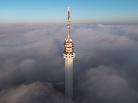 Aerial view of the Zendmast Roermond telecommunications tower rising above a thick layer of low clouds and fog under a clear blue sky in Roermond, Limburg, Netherlands.