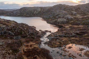 Fototapeta premium Drone view of a small stream flowing over rocks into a tranquil bay of the Arctic Ocean. People hike on the rugged coastal tundra of the Murmansk Region at sunset.