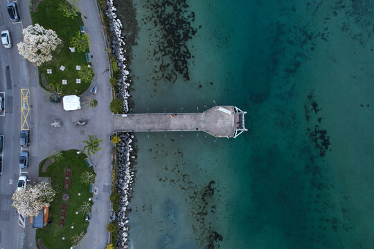 Aerial view of a concrete pier extending into the turquoise waters of Lake Geneva with blooming trees and a green park in Morges, Vaud, Switzerland.