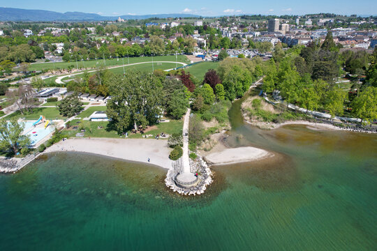 Aerial view of the scenic lakefront featuring a sandy beach, lush green parkland, and the river mouth flowing into the clear turquoise waters of Lake Geneva in Morges, Vaud, Switzerland.