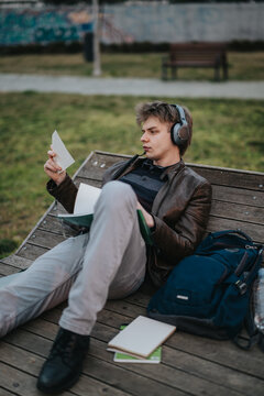 A high school student wearing headphones reads loose notes while reclining on a wooden park bench. The teenage student studies with notebooks and a backpack beside him in an outdoor setting.