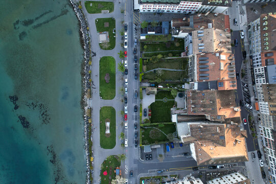 Aerial view of the waterfront promenade with green parks, parked cars along the street, and historic rooftops by the lake in Morges, Vaud, Switzerland.