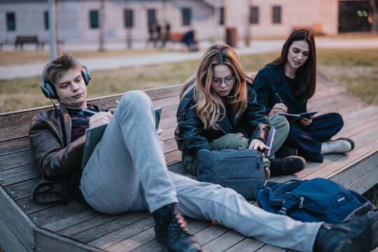High school students sit on a wooden bench studying and writing in notebooks while one listens to music. Three friends relax outdoors with backpacks and journals during a casual study session.