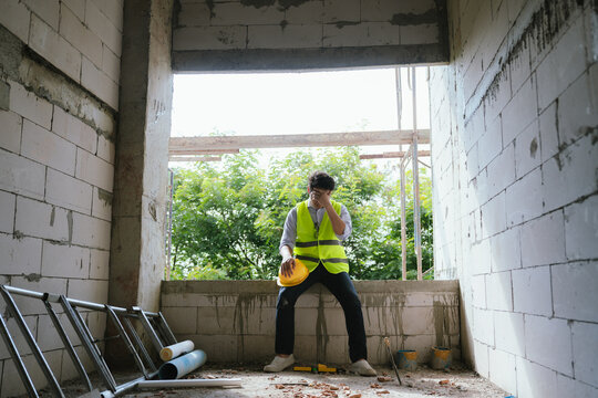 Tired construction worker resting at a building site while holding a safety helmet, representing workplace stress, fatigue, and construction industry challenges.