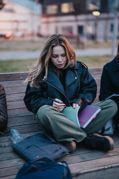 A high school student sits on a wooden bench writing in a notebook while concentrating. A young teen studies outdoors with a pen and open notebook on her lap in a casual setting.
