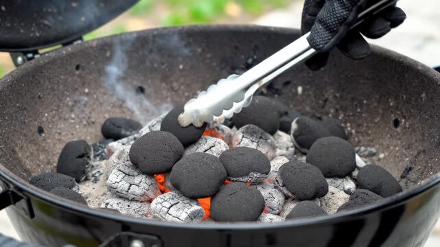 A person using metal tongs to rearrange hot glowing charcoal briquettes in a black outdoor barbecue grill.