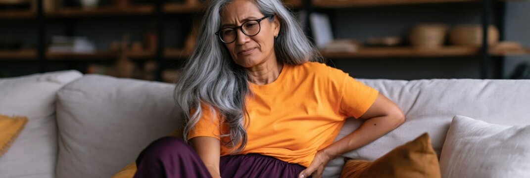 Mature woman grimacing and holding lower back on sofa in living room