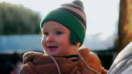 Baby smiling in bear teddy fleece coat and green beanie hat while held outdoors, joyful infant enjoying winter day in parent’s arms with warm family connection and happiness © Marco
