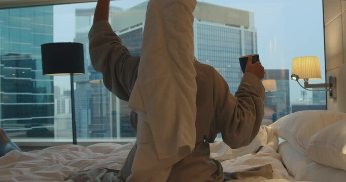 Woman wearing bathrobe and towel dancing joyfully while sitting on a bed in a luxury hotel room. City skyline view through window in the morning. Back view medium shot.