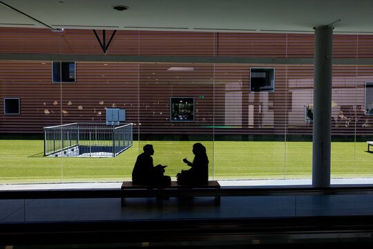 Two business silhouettes seated on a bench before a conference meeting, minimalist corporate setting with courtyard view, clean architectural lines, professional and modern atmosphere.