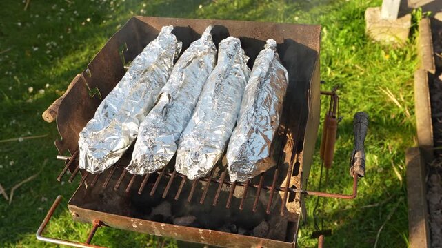 Fish wrapped in foil cooking on barbecue grill outdoors. Open fire meal preparation, camping food, picnic, grilling process and summer outdoor cooking concept.