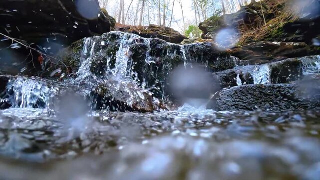 Close up of water splashing over mossy mountain rocks. Low angle shot of a stream cascading over dark wet stones with water drops on lens.