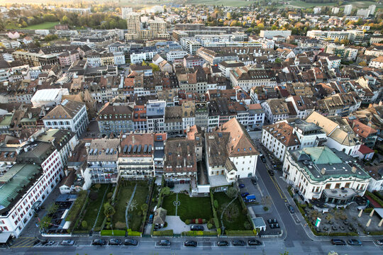 Aerial view of the historic town center with its traditional red-tiled rooftops, narrow streets, and lush green gardens under soft evening light in Morges, Vaud, Switzerland.