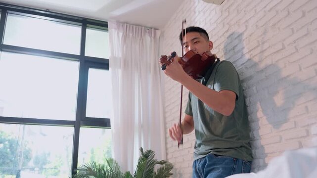 Asian young men Leaning against the wall and practicing violin, they skilled the strings to produce melodious tunes, artistic lifestyle playing violin in the living room for practice  and  hobby