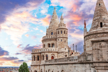 Fototapeta premium The Fisherman's Bastion in Budapest stands tall against a vibrant sunset sky filled with colorful clouds.