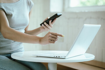 Fototapeta premium Woman using a smartphone beside a laptop and calculator at a table, representing online banking, remote work, digital communication, and modern lifestyle.