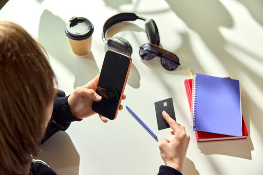 Teenager holding smartphone near bank card and notebooks on bright table. Concept of student budgeting, online payment, digital learning, lifestyle planning and youth finance.