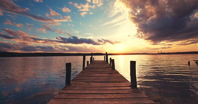 Wooden pier leading towards a vibrant sunset horizon with scenic clouds over calm lake water