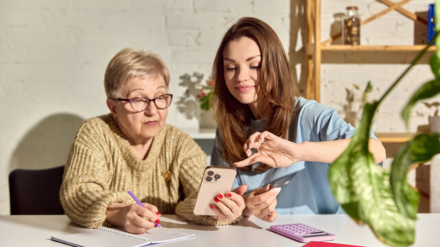 Woman helping senior mother with mobile money transfer at home. Concept of family finance, online banking, elder support, fintech education and secure digital payment.