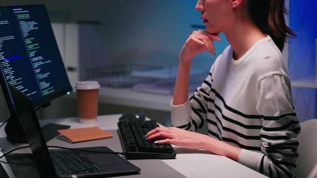 Focused Asian woman coding on computer during overtime night shift in modern office. Software engineering, startup workflow, remote development and women in technology concept.