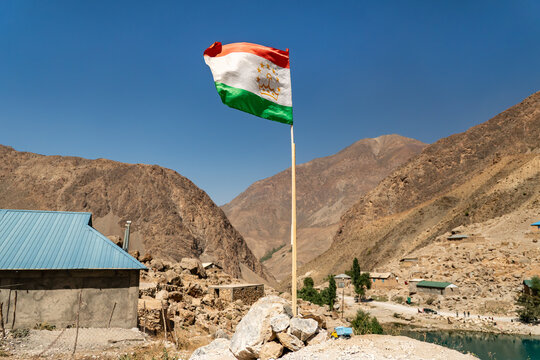 Flag of Tajikistan waving against blue sky with mountains and a mountain village aul in the background.