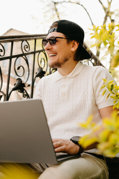 Young Man Working On Laptop On Sunny Balcony Wearing Backward Cap And Sunglasses Smiling While Checking Screen