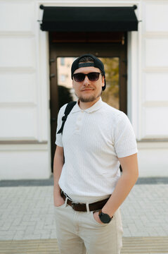 Man Smiling With Backpack Urban Walks Past White Storefront In Casual White Polo, Friendly Approachable