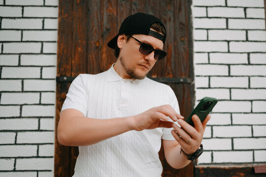 Caucasian Man On Street Checking Smartphone, Wearing White Tee And Backward Cap, Sunglasses, Leaning