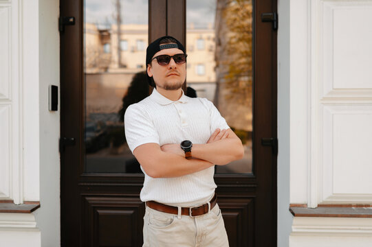 Man With Cap Arms Crossed Poses Assertive In Front Of Dark Entrance, Wearing White Polo And Sunglasses,
