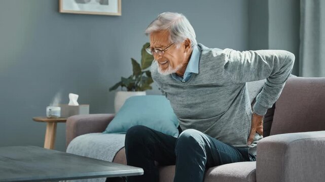 Elderly man sitting on couch grimacing in pain clutching lower back discomfort at home