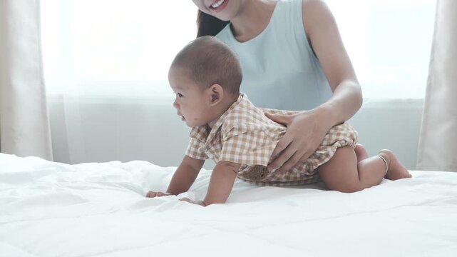 A seven-month-old baby boy, wearing a cute brown outfit and a bear hat, plays with a colorful ball with his Asian mother, who holds him on the bed in their bedroom at home.
