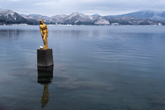 JAPAN - DEC 25, 2024: Tatsuko Golden Statue at Lake Tazawa in Akita Japan with snowy mountains background, iconic landmark reflecting on calm water, winter travel destination landscape and famous spot