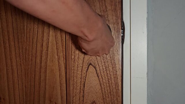 A hand reaches out to turn a round stainless steel doorknob on a modern light-brown wooden door