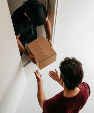 Delivery courier in black uniform handing sealed cardboard package to customer receiving at home doorway showing fast online shopping home delivery service concept