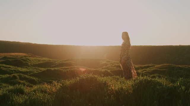 Woman In Sunlit Meadow At Sunset, Backlit Silhouette In Long Flowing Dress, Rolling Hills And Tall Grass