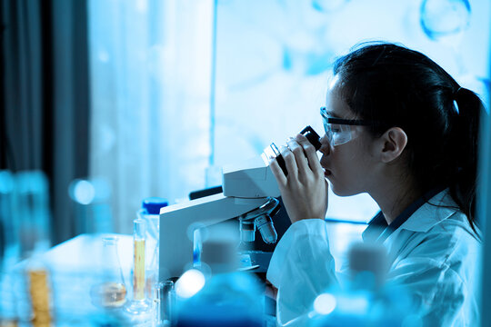 A woman wearing a lab coat and safety glasses is looking through a microscope. She is focused on the image in front of her, likely studying something related to science or research