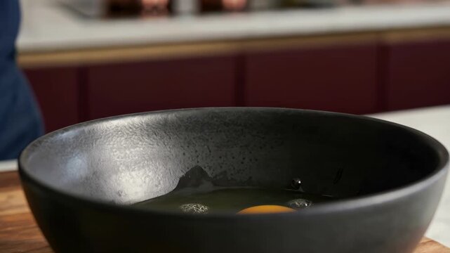 Close up footage of a chef cracking a fresh egg into a black ceramic bowl in a professional kitchen environment with intentional movement and high-quality culinary presentation.