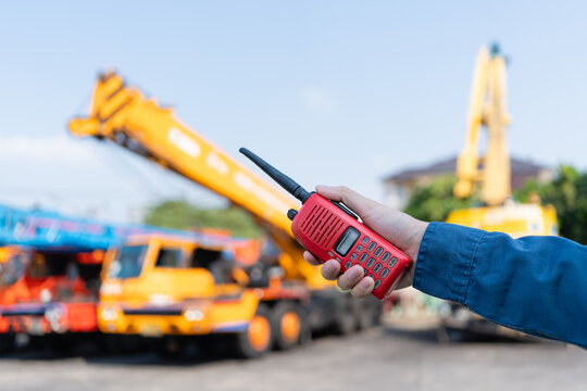 Worker hand holds a two way radio for crane safety communication at a heavy equipment yard, coordinating lifting operations, inspection tasks, and onsite risk control.
