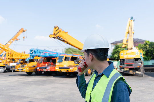 Engineer uses a radio beside mobile cranes at a heavy equipment yard, coordinating crane inspection, safety communication, lifting readiness, and onsite risk control.