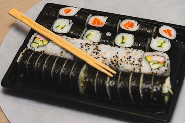 Large assorted sushi set in a black takeaway tray with wooden chopsticks on top. Concept of Japanese food delivery, healthy takeout meal, and oriental cuisine variety. Top view of maki rolls, uramaki, © Anna Kondratiuk
