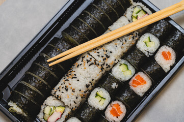 Large assorted sushi set in a black takeaway tray with wooden chopsticks on top. Concept of Japanese food delivery, healthy takeout meal, and oriental cuisine variety. Top view of maki rolls, uramaki, © Anna Kondratiuk